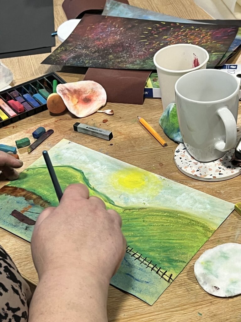 A participant working on a pastel drawing of a hill with a yellow sun above